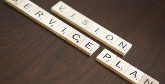 A picture of Scrabble tiles on a brown table spelling out 'Vision', 'Service', and 'Plan'
